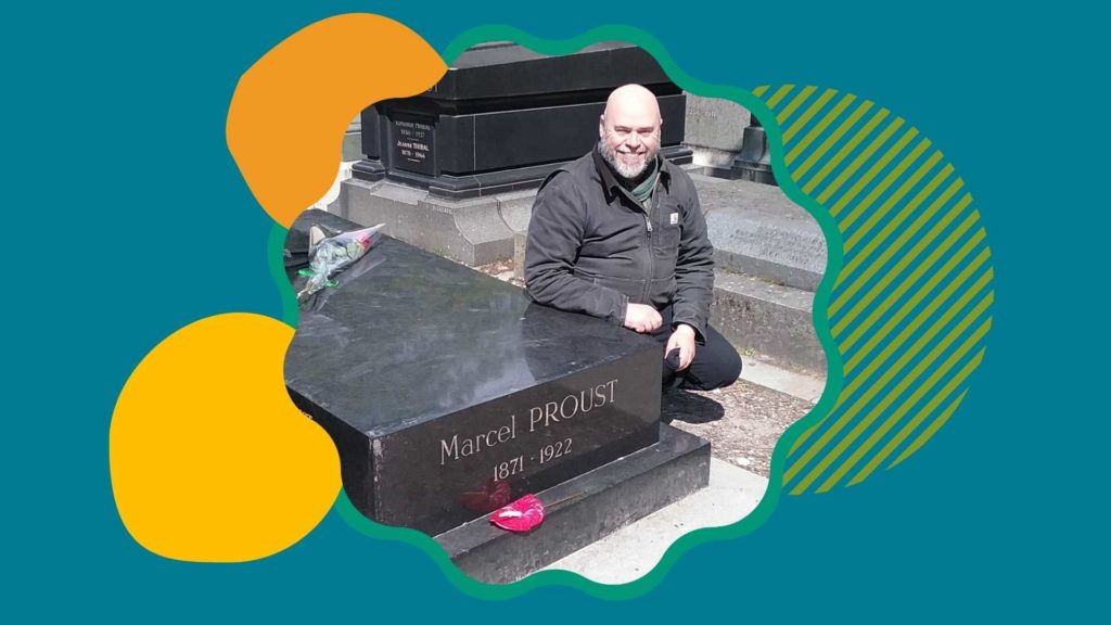 A smiling Drew Bourn in front the grave of Marcel Proust, in a circular frame with orange, yellow, and green decorative circles behind the photo on a teal background