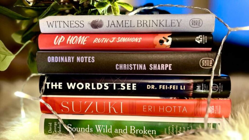 Stack of hardcover books, from the top down: Witness by Jamel Brinkley, Up Home by Ruth J Simmons, Ordinary Notes by Christina Sharpe, The Worlds I See by D. Fei-Fei Li, Suzuki by Eri Hotta, and Sounds Wild and Broken by David George Haskell. Below the books is a white furry blanket and a strand of fairy lights surround the book stack.