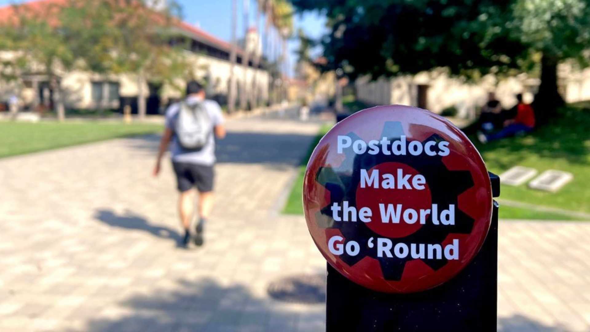 photo of button that says "Postdocs Make the World Go 'Round" with Stanford Campus in the background